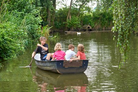 Vier Kinder rudern in einem kleinen Boot auf einem ruhigen Teich, umgeben von dichter Vegetation und Bäumen.