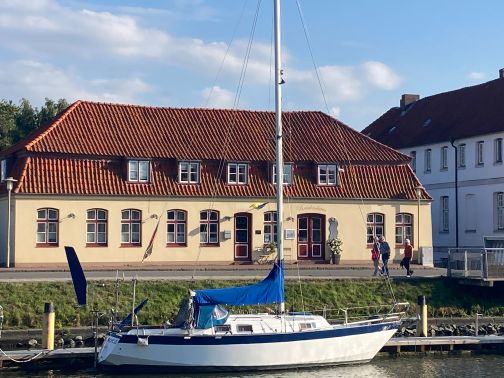 Das Brückenhaus am Hafen mit rotem Ziegeldach, Segelboot im Vordergrund und Spaziergängern am Steg.