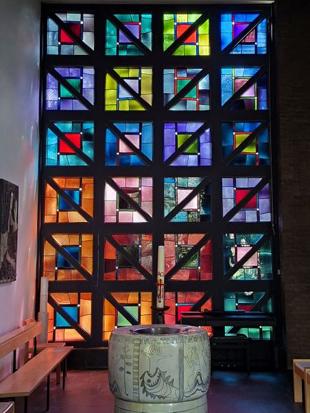 Buntes Kirchenfenster mit geometrischen Mustern. Ein Taufbecken mit Kerze steht davor. 