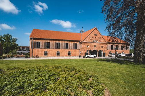 Hotel Breitenburg vor grüner Wiese, ein historisches Backsteingebäude mit rotem Dach und geparkten Autos bei blauem Himmel.
