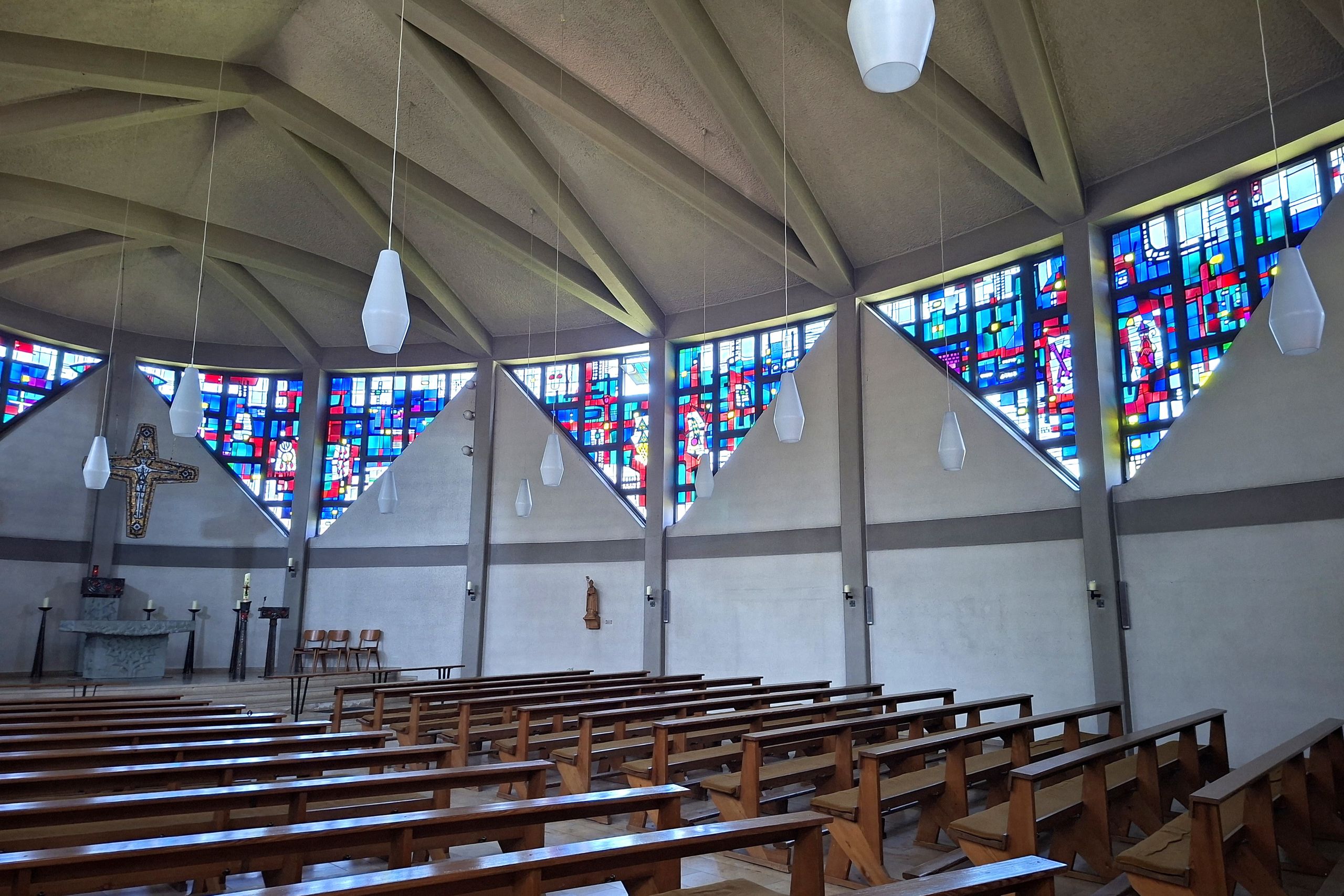 Innenansicht der Kirche St. Vicelin in Oldenburg. Bunte Glasfenster und Kirchenbänke aus Holz prägen den Raum. Moderne Architektur mit hängenden Lampen.