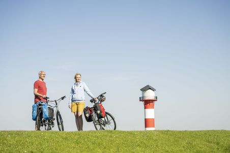 Zwei Personen mit Fahrrädern stehen auf einem grünen Deich. Im Hintergrund ist ein rot-weiß gestreifter Leuchtturm zu sehen.