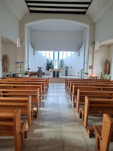 Moderne Kirche mit Holzbänken und einem schlichten Altar. Große Fenster im Hintergrund lassen viel Licht herein.