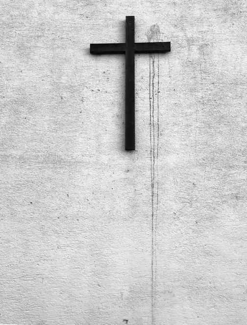 Ein schwarzes Kreuz hängt an einer weißen, strukturierten Wand. Die Wand zeigt Spuren von Verwitterung.