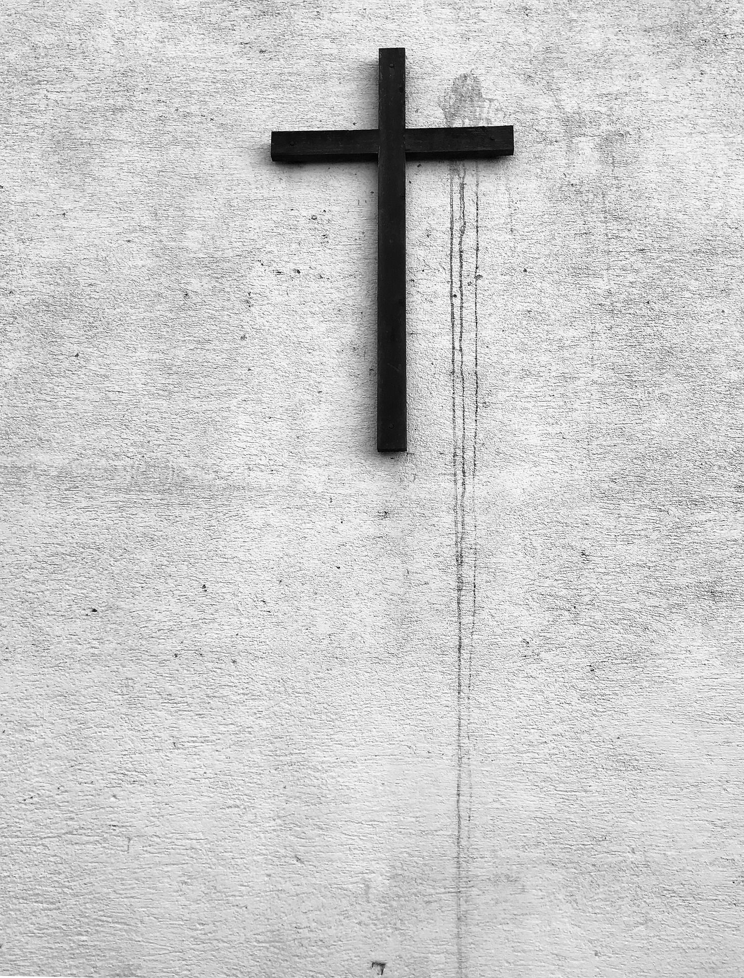 Ein schwarzes Kreuz hängt an einer weißen, strukturierten Wand. Die Wand zeigt Spuren von Verwitterung.