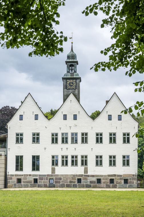 Historisches Gebäude mit Giebeldach und Uhrturm, umrahmt von grünen Bäumen. Der Himmel ist bewölkt.