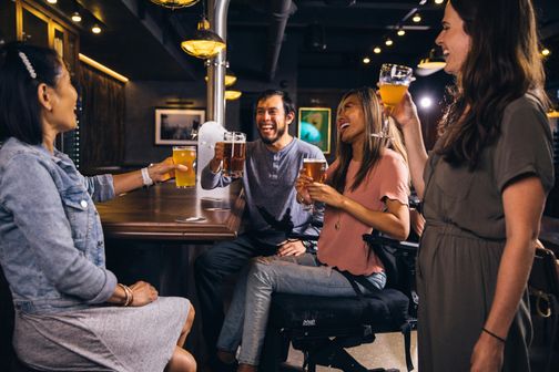Vier Personen sitzen in einer Bar und stoßen mit Biergläsern an. Sie lachen und scheinen eine gute Zeit zu haben.