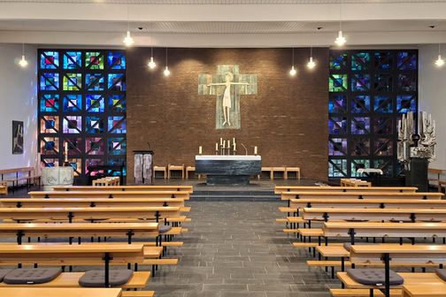 Innenansicht der Kirche St. Josef mit bunten Glasfenstern, einem schlichten Altar und Holzbänken. Die Beleuchtung ist dezent und warm.