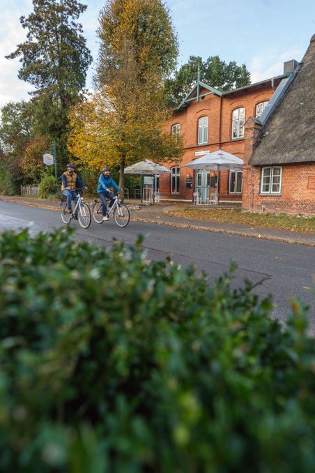Zwei Radfahrer fahren an einem Backsteingebäude mit Reetdach vorbei. Herbstliche Bäume und ein Café mit Sonnenschirmen sind im Hintergrund zu sehen.
