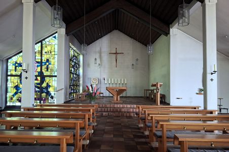 Innenansicht der St. Franziskus Xaverius-Kirche auf Fehmarn. Holzbänke führen zum Altar mit einem Kreuz. Bunte Glasfenster zieren die Seitenwände.