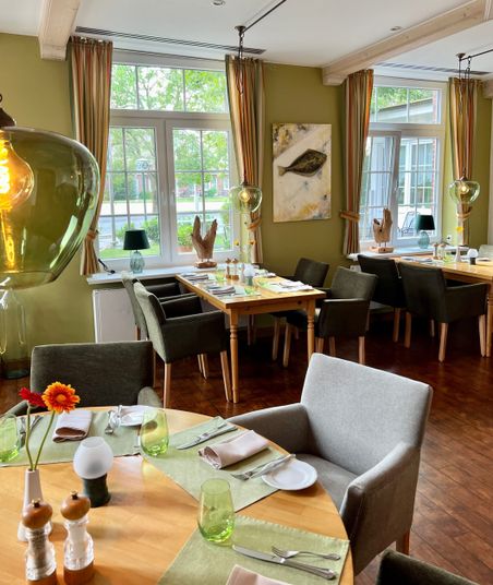 Ein stilvoll eingerichtetes Restaurant mit gedeckten Tischen, grünen Wänden und großen Fenstern. Kunstwerke und Dekorationen schaffen eine einladende Atmosphäre.
