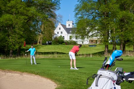 Zwei Golfer auf einem grünen Rasen vor einem weißen Schloss. Einer puttet, während der andere die Fahne hält. Im Vordergrund ein Golfbag.
