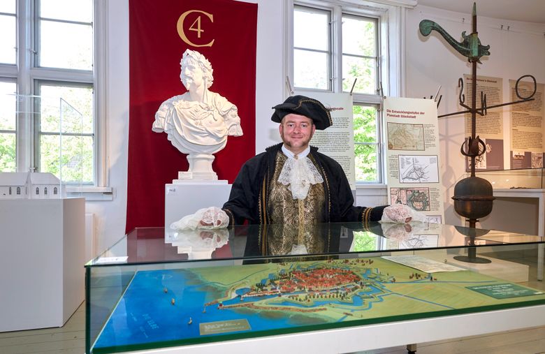 Ein Mann in historischer Kleidung steht in einem Museum vor einer Büste und einer Karte. Im Hintergrund sind informative Tafeln und ein roter Vorhang zu sehen.