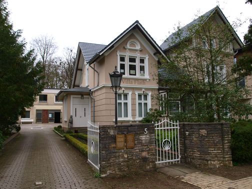 Die Villa Flath mit gepflegtem Garten und weißem Tor. Ein Weg führt zur Eingangstür, flankiert von Bäumen und einem modernen Anbau im Hintergrund.