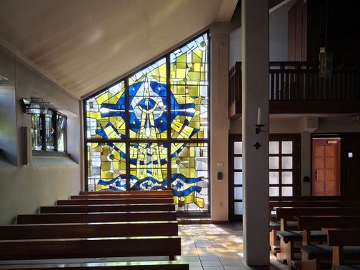 Blick in die St. Franziskus Xaverius-Kirche auf Fehmarn. Ein großes, buntes Glasfenster dominiert den Raum, umgeben von Holzbänken.