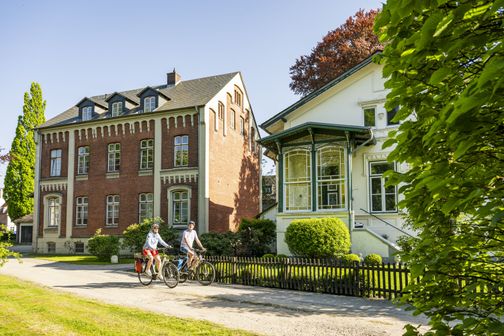 Zwei Radfahrer fahren an einem sonnigen Tag an zwei historischen Gebäuden in Itzehoe vorbei. Das linke Gebäude ist aus rotem Backstein, das rechte ist weiß.