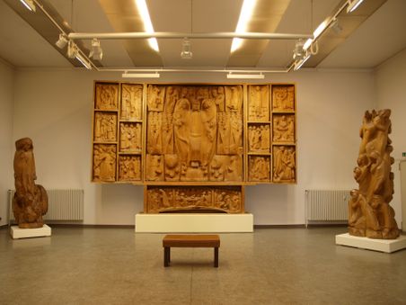 Ein Ausstellungsraum der Kunsthalle Otto Flath mit einem großen Holzrelief an der Wand und zwei Holzstatuen auf Sockeln. Ein Holzhocker steht in der Mitte.