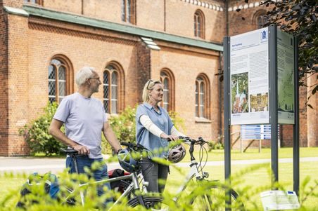 Zwei Radfahrer stehen vor einer Informationstafel des Mönchswegs auf dem Kirchplatz in Bad Segeberg. Im Hintergrund ist eine Backsteinkirche zu sehen.