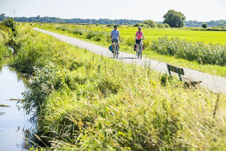 Zwei Radfahrer fahren auf Mönchsweg durch die grünen Wümmewiesen bei Bremen. Ein kleiner Wasserlauf und eine Bank sind zu sehen.