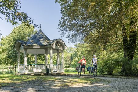 Zwei Radfahrer mit Helmen stehen neben einem Pavillon im Bürgerpark Bremen, umgeben von Bäumen und sonnigem Wetter.