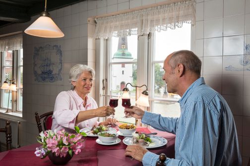 Ein älteres Paar genießt ein Essen in einem gemütlichen Restaurant und stößt mit Rotwein an. Durch das Fenster sieht man einen Turm.