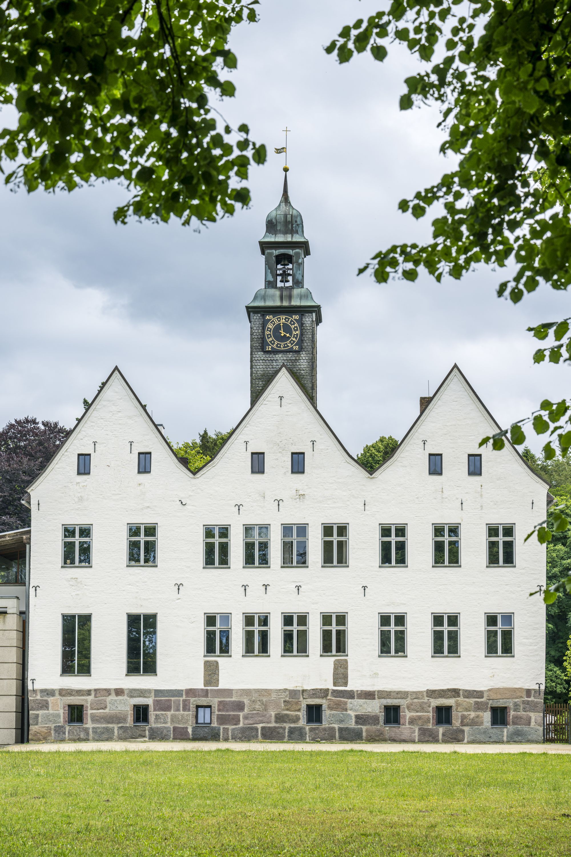 Historisches Gebäude mit Giebeldach und Uhrturm, umrahmt von grünen Bäumen. Der Himmel ist bewölkt.