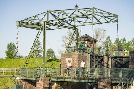 Zwei Radfahrer stehen auf einer historischen Schleusenbrücke in Kasenort. Die Brücke ist aus Metall und Ziegelstein, umgeben von grüner Landschaft.