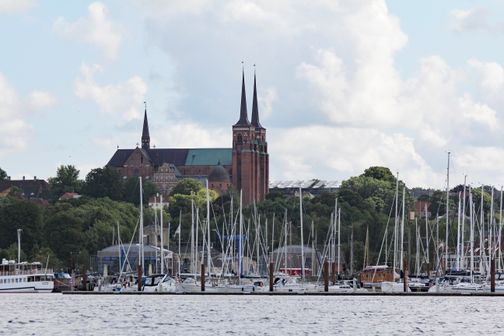 Der Dom zu Roskilde erhebt sich majestätisch über den Hafen, umgeben von Segelbooten und üppigem Grün unter einem bewölkten Himmel.
