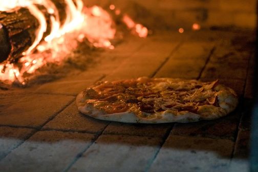 Eine Pizza wird in einem traditionellen Holzofen gebacken. Der Ofen ist mit brennendem Holz beheizt, das eine warme, einladende Atmosphäre schafft.