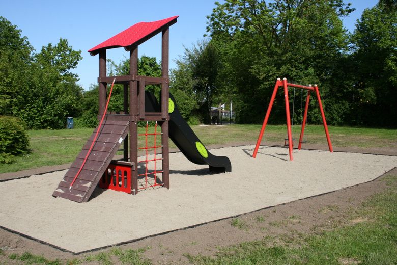 Ein Spielplatz mit Klettergerüst, Rutsche und Schaukel auf einer Sandfläche, umgeben von grünen Bäumen und Rasen.