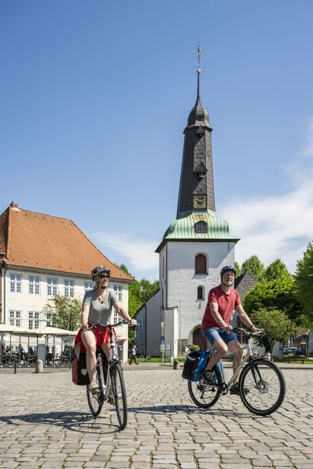 Zwei Radfahrer fahren auf einem gepflasterten Platz vor der Kirche in Glückstadt. Der Himmel ist klar und blau, die Umgebung grün und einladend.