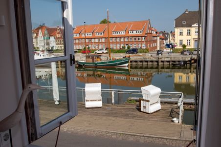 Blick durch ein offenes Fenster auf den Hafenkanal mit Segelboot und roten Backsteinhäusern; die Szene spiegelt sich im Wasser.