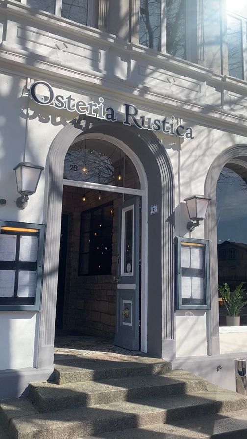 Eingang der Osteria Rustica mit offener Tür, Treppe und Laternen. Die Fassade ist weiß und das Schild zeigt den Restaurantnamen.