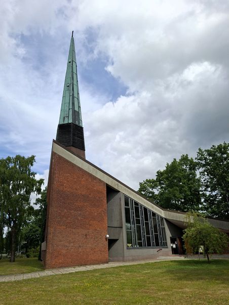 Die Friedenskirche Trappenkamp zeigt eine moderne Architektur mit spitzem Turm und Backsteinfassade. Sie ist von Bäumen und einer Wiese umgeben.