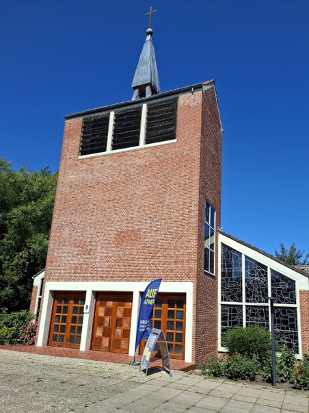 Die St. Franziskus Xaverius-Kirche auf Fehmarn zeigt einen Backsteinturm mit Kreuz und moderne Glasfenster. Ein Banner steht vor dem Eingang.