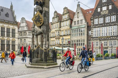 Radfahrer passieren den Bremer Marktplatz mit dem Roland im Hintergrund. Historische Gebäude und Passanten beleben die Szene.