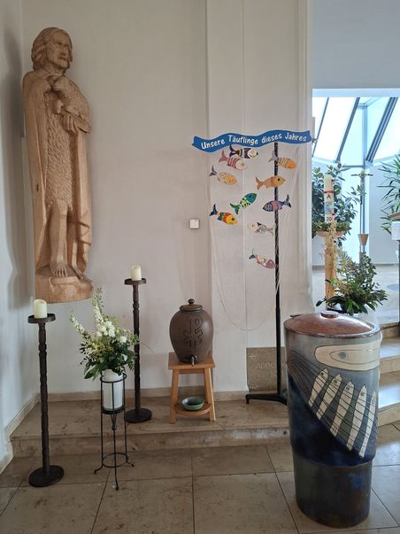 Kircheninnenraum mit Taufbrunnen, einer Holzstatue, Kerzenständern und Dekoration für Täuflinge. Ein Schild zeigt "Unsere Täuflinge dieses Jahres".