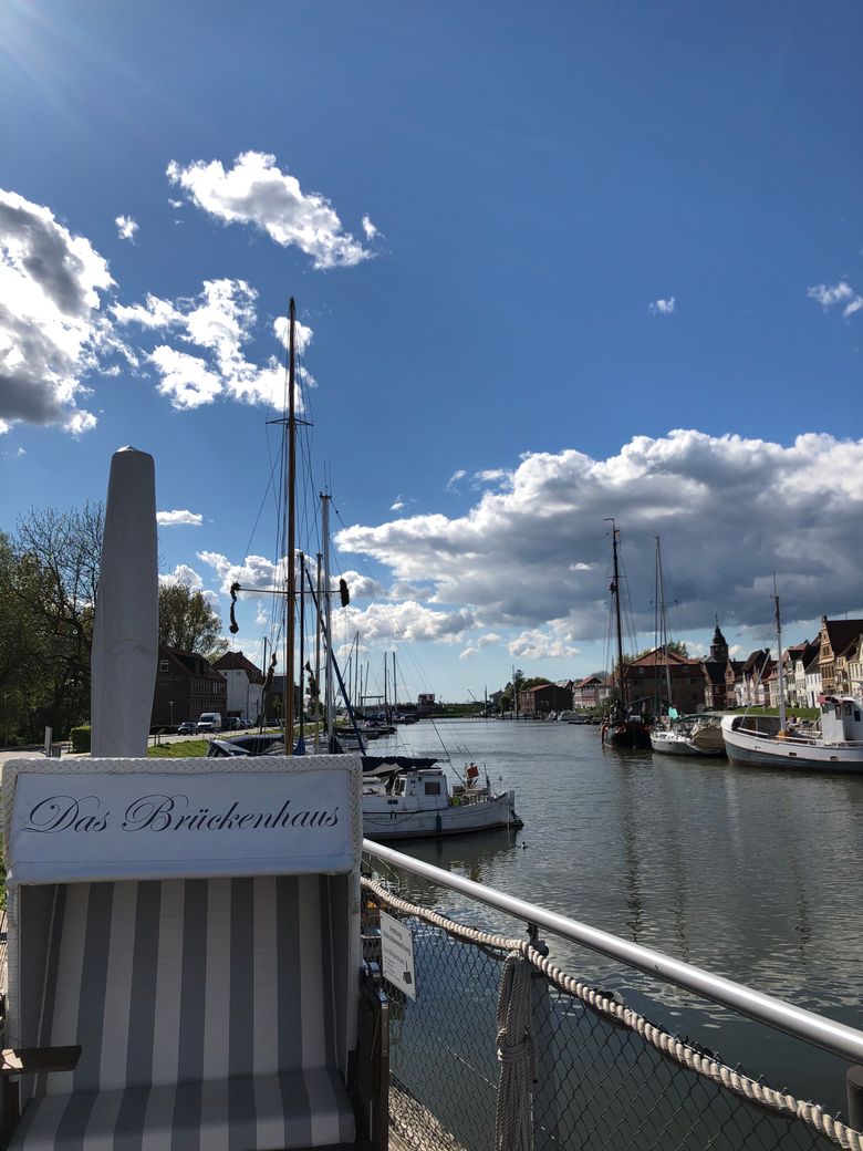 Hafenansicht mit Segelbooten, Wasser und Häusern am Ufer; im Vordergrund das Brückenhaus-Schild unter blauem Himmel mit Wolken.