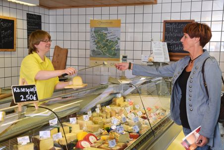 Eine Verkäuferin in gelbem Hemd reicht einer Kundin ein Stück Käse über die Theke. Verschiedene Käsesorten sind ausgestellt.