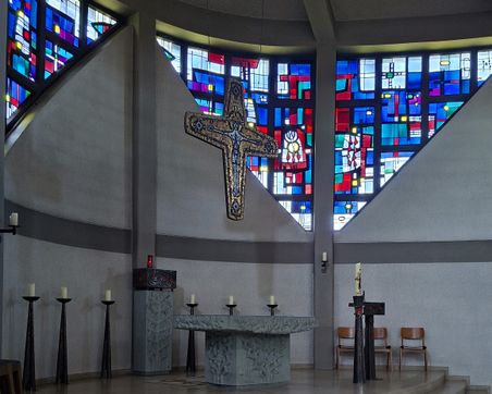 Innenansicht der Kirche St. Vicelin in Oldenburg. Bunte Glasfenster und ein großes Kreuz dominieren den Raum über dem Altar.